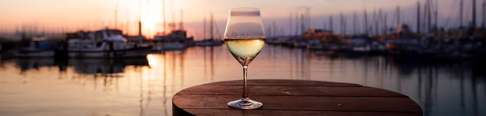 Weißweinglas auf Holztisch vor einem Hafen.