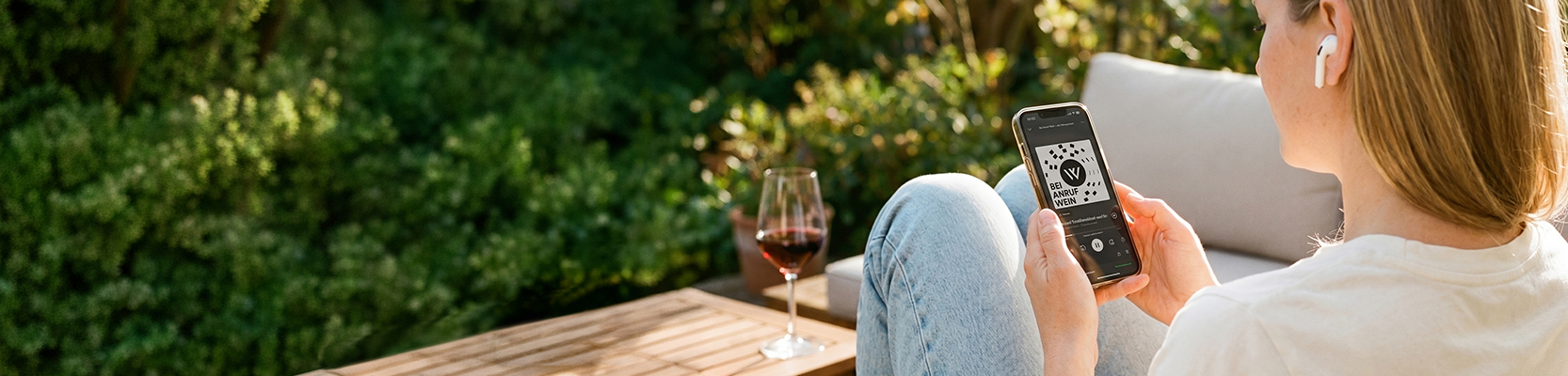 Eine Person sitzt im Garten mit einem Glas Wein und hört den Podcast Bei Anruf Wein.